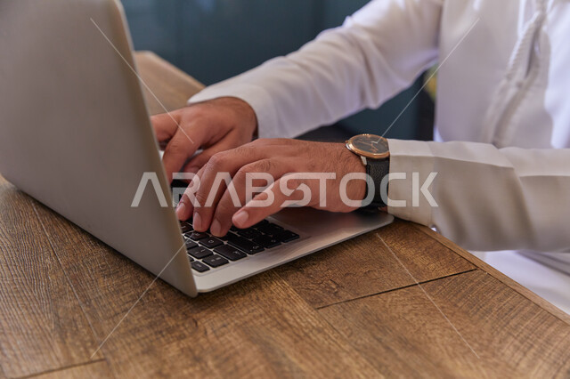 Chats and conversations with friends. Browsing social media and following social media news. A close-up of an Emirati Arab man wearing a wristwatch working on a computer, doing online work from home.