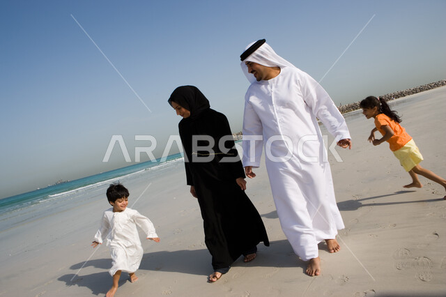 أخوان يلعبان لعبة المطاردة ويجريان حول والديهم, السياحة في دول الخليج العربي والاستمتاع بالعطلة الصيفية مع العائلة، قضاء أوقات الإجازة مع الأبناء, مفهوم الاسرة الإماراتية السعيدة، أب وأم عربيان خليجيان إماراتيان يقومان باللعب مع أبنائهم على شاطئ البحر
