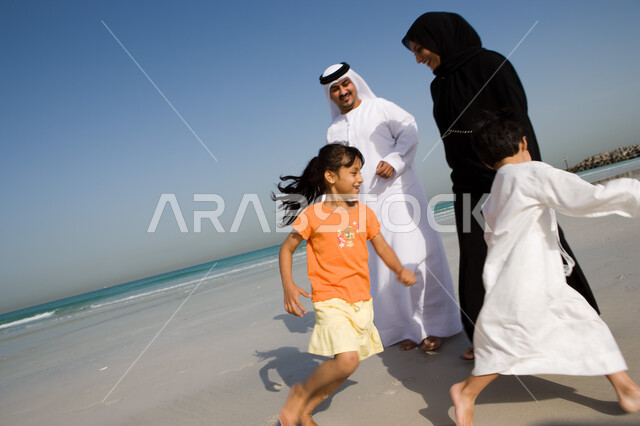أخوان يلعبان لعبة المطاردة ويجريان حول والديهم, السياحة في دول الخليج العربي والاستمتاع بالعطلة الصيفية مع العائلة، قضاء أوقات الإجازة مع الأبناء, مفهوم الاسرة الإماراتية السعيدة، أب وأم عربيان خليجيان إماراتيان يقومان باللعب مع أبنائهم على شاطئ البحر