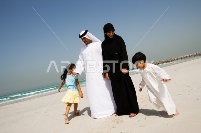 أخوان يلعبان لعبة المطاردة ويجريان حول والديهم, السياحة في دول الخليج العربي والاستمتاع بالعطلة الصيفية مع العائلة، قضاء أوقات الإجازة مع الأبناء, مفهوم الاسرة الإماراتية السعيدة، أب وأم عربيان خليجيان إماراتيان يقومان باللعب مع أبنائهم على شاطئ البحر