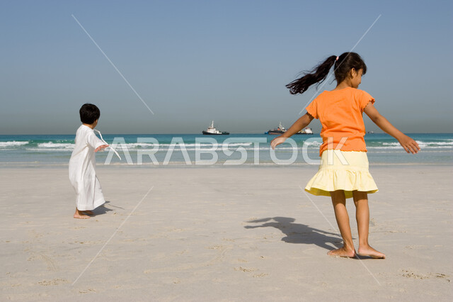 الإستمتاع بالفعاليات الصيفية الممتعة مع الأخوة, فتاة صغيرة عربية إمراتية خليجية تقوم باللعب على شاطئ البحر رفقة أخيها في رحلة صيفية, أنشطة ترفيهية عائلية في العطلة, منتجعات شاطئية في الإمارات