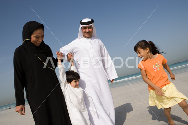 مفهوم الأسرة السعيدة وحب الآباء للأبناء, الاستمتاع بالعطلة الصيفية مع العائلة، قضاء أوقات الإجازة مع الأبناء,  أب وأم عربيان خليجيان إماراتيان يمسكان يد ابنهم على شاطئ البحر، منتجعات شاطئية إماراتية