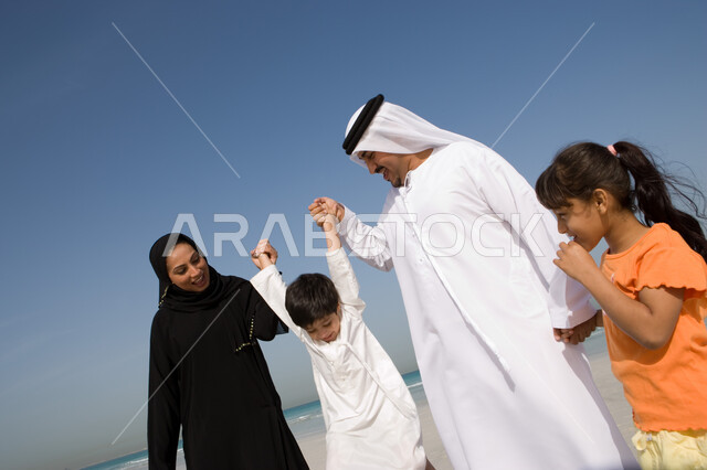 مفهوم الأسرة السعيدة وحب الآباء للأبناء, الاستمتاع بالعطلة الصيفية مع العائلة، قضاء أوقات الإجازة مع الأبناء,  أب وأم عربيان خليجيان إماراتيان يمسكان يد ابنهم على شاطئ البحر، منتجعات شاطئية إماراتية