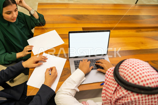 اجتماع اداري مع مجموعة من الأشخاص السعوديين الخليجيين في غرفة الاجتماعات  ، اجتماع رجال اعمال و سيده اعمال سويا داخل الشركة ، استخدام الكمبيوتر المحمول لادارة الاعمال ،  شركة سعودية ، عمل خليجي ،  فريق العمل 