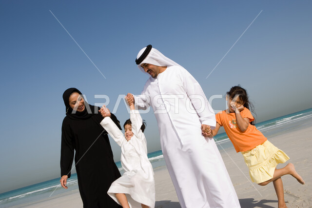 الاستمتاع بالعطلة الصيفية مع العائلة، قضاء أوقات الإجازة مع الأبناء, مفهوم الاسرة الإماراتية السعيدة، أب وأم عربيان خليجيان إماراتيان يمسكان يد ابنائهم ويقومان باللعب معهم على شاطئ البحر، منتجعات شاطئية إماراتية