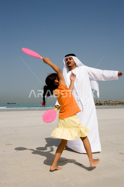 The concept of fatherhood and the participation of children in their activities, entertainment and summer family activities, family entertainment activities on vacation, an Arab Emirati father wearing a ghutra and an Emirati kandora playing tennis with hi