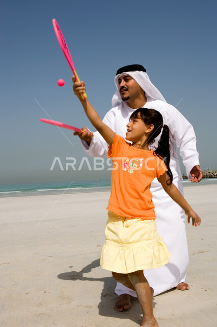 The concept of fatherhood and the participation of children in their activities, entertainment and summer family activities, family entertainment activities on vacation, an Arab Emirati father wearing a ghutra and an Emirati kandora playing tennis with hi