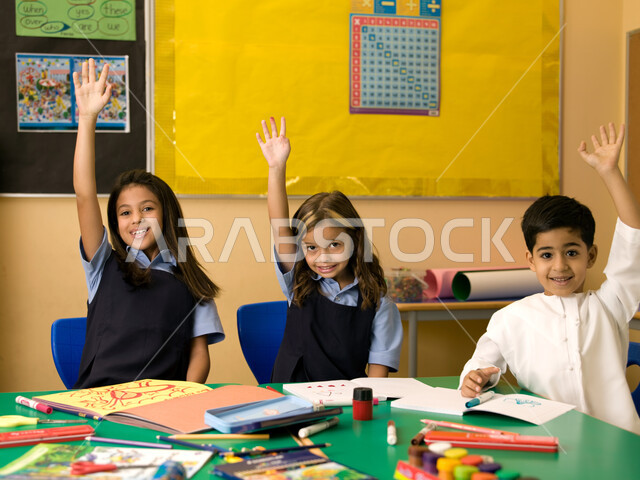 Enthusiasm, interaction and participation with the teacher by raising the hand, commitment and discipline to the rules of the school classroom, stationery and drawing and coloring tools, Emirati Gulf Arab students wearing Emirati school uniforms raising t