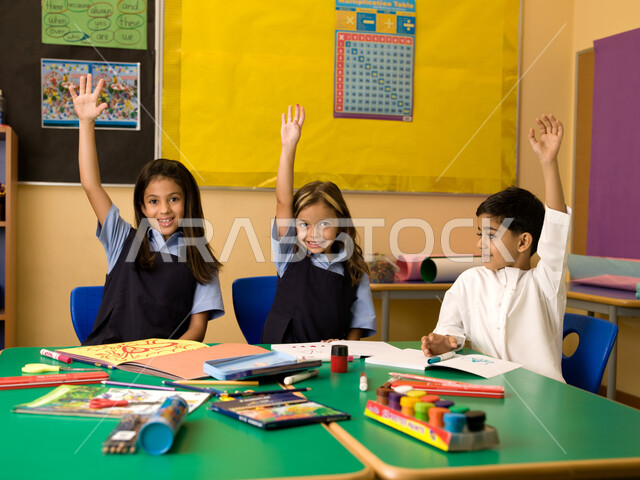 الحماس والتفاعل والمشاركة مع المعلمة برفع اليد، الالتزام والانضباط بقواعد الفصل المدرسي، قرطاسية وأدوات الرسم والتلوين، تلاميذ عرب خليجيون إماراتيون يرتدون الزي المدرسي الإماراتي يرفعون أيديهم عاليا.