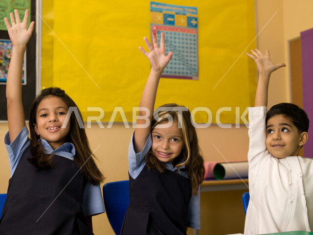 الحماس والتفاعل والمشاركة مع المعلمة برفع اليد، الالتزام والانضباط بقواعد الفصل المدرسي، صورة مقربة لتلاميذ عرب خليجيين إماراتيين يرتدون الزي المدرسي الإماراتي يرفعون أيديهم عاليا، المدارس التعليمية في الإمارات العربية المتحدة.