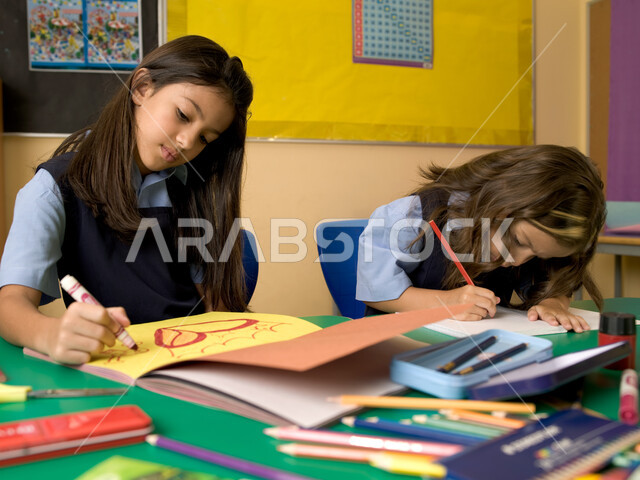 الاندماج والتركيز في الأنشطة الفنية، قرطاسية وأدوات التلوين، طالبتان عربيتان خليجيتان إماراتيتان ترتديان الزي المدرسي وتقومان بالرسم في الفصل المدرسي، المدارس التعليمية في الإمارات العربية المتحدة.