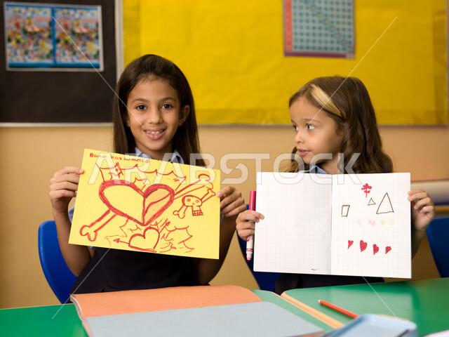 التعبير بالرسم عن مشاعر الحب والمودة، القرطاسية وأدوات التلوين، طالبتان عربيتان خليجيتان إماراتيتان ترتديان الزي المدرسي وتقومان بعرض الرسومات، المدارس التعليمية في الإمارات العربية المتحدة، ايماءات تدل على السعادة والفرح بالإنجاز.