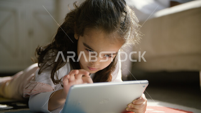 الاستمتاع بالألعاب الترفيهية على الأجهزة الذكية, صورة مقربة لفتاة عربية خليجية إماراتية تجلس في غرفة المعيشة وتستخدم التابليت, استخدام التقنيات والتطبيقات التعليمية الحديثة لدى الأبناء, مفهوم التسلية والترفيه وقضاء وقت ممتع