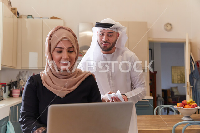 توصية طلبيات عبر الانترنت من المنزل، قضاء أوقات ممتعة بالعمل الأونلاين، زوجان عربيان خليجيان يقومان بتصفح مواقع التواصل الاجتماعي في المطبخ، إنجاز الأعمال المطلوبة عبر جهاز الحاسوب، متابعة أحدث الأخبار في السوشل ميديا.