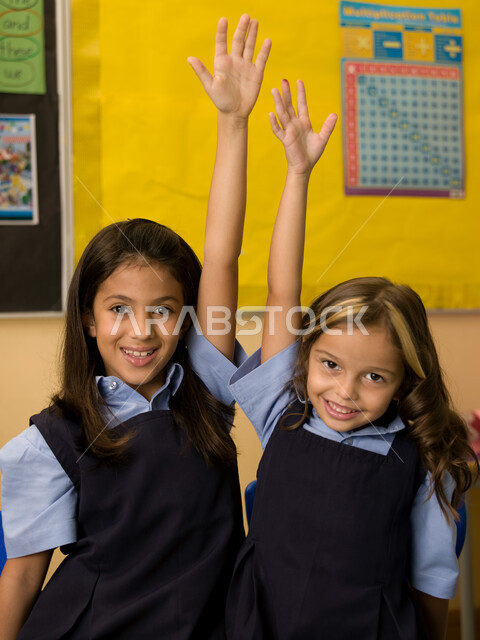 التفاعل والمشاركة مع المعلمة برفع اليد،  طالبتان عربيتان خليجيتان إماراتيتان ترتديان الزي المدرسي ترفعان ايديهما، المدارس التعليمية في الإمارات العربية المتحدة، ايماءات تدل على السعادة والفرح بالإنجاز، الالتزام والانضباط بقواعد الفصل المدرسي.