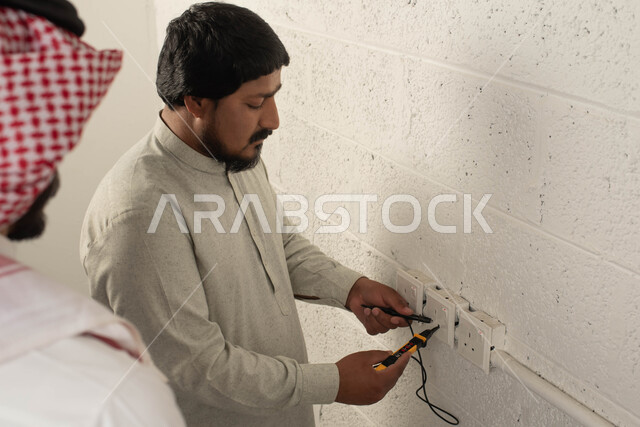 إصلاح الأعطال المنزليه, فحص مفتاح الكهرباء, عامل صيانه يستخدم جهاز قياس التيار الكهربائي (الأميتير), صورة مقربة من الخلف لشاب عربي خليجي سعودي يرتدي الثوب التقليدي والشماغ ينظر إلى شيء ما, أدوات ومعدات الصيانه الكهربائية