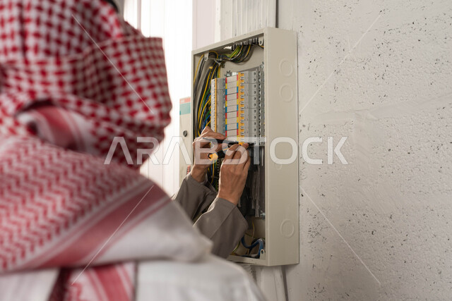 صيانة علبة الأسلاك الكهربائية, تركيب لوحة عداد الكهرباء, صورة مقربة من الخلف لرجل عربي خليجي سعودي يرتدي الشماغ التقليدي يقف بجانب المهندس, عامل فني يستخدم جهاز قياس التيار الكهربائي (الأميتر), أدوات ومعدات الفنيين, استقدام العمالة من الخارج