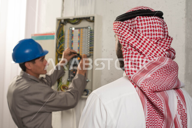 أدوات ومعدات الفنيين, صيانة علبة الأسلاك الكهربائية, تركيب لوحة عداد الكهرباء, صورة مقربة من الخلف لرجل عربي خليجي سعودي يرتدي الشماغ التقليدي يقف بجانب المهندس, عامل فني يستخدم جهاز قياس التيار الكهربائي (الأميتر), استقدام العمالة من الخارج