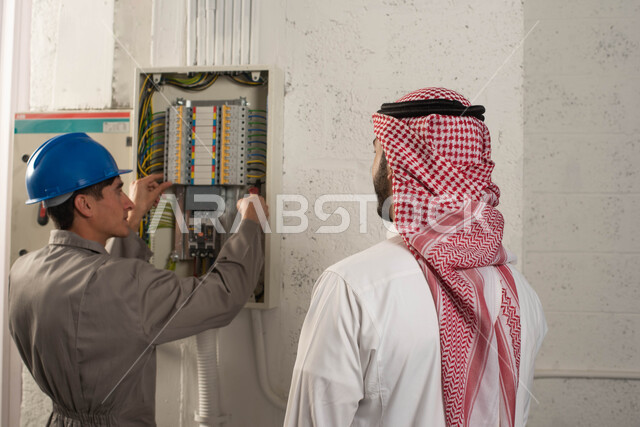 صيانة وتركيب لوحة عداد الكهرباء, مفهوم الهندسة الكهربائية, عامل فني كهربائي يستخدم مفك فحص الكهرباء, صورة مقربة من الخلف لرجل عربي خليجي سعودي يرتدي الشماغ والثوب التقليدي يقف بجانب المهندس, ارتداء ملابس العمل وخوذه الحماية