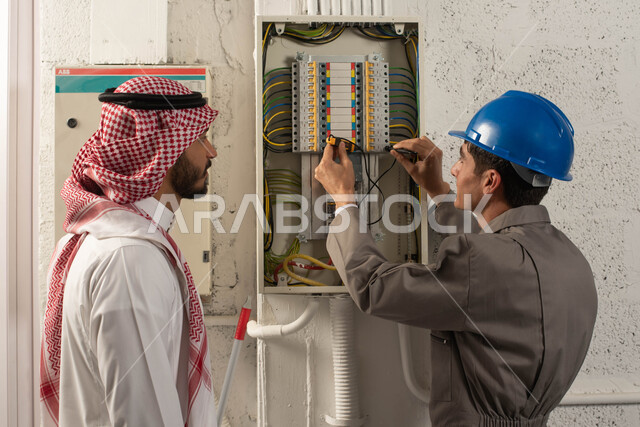 A technical worker uses an ammeter, a close-up picture from the back of a Saudi Gulf Arab man wearing a shemagh and traditional thobe, standing next to the engineer, wearing work clothes and a protective helmet, maintenance and installation of the electricity meter panel, electrical engineering concept