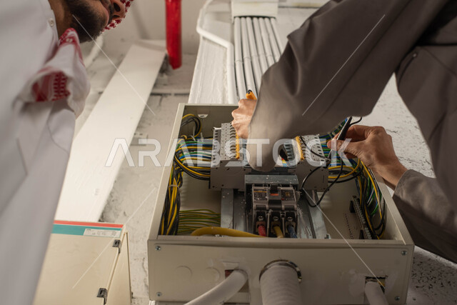 Electrical engineering concept, close-up image of a technician’s hand using an ammeter, a Saudi Gulf Arab man wearing a thobe and shemagh, maintenance and installation of the electricity meter panel, installing switches and extending wires, working in the engineering field, bringing in workers from abroad