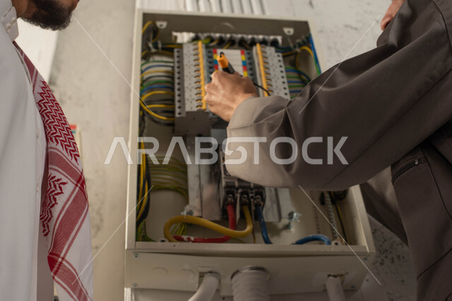 تركيب المفاتيح وتمديد الأسلاك, مفهوم الهندسة الكهربائية, صورة مقربة ليد عامل فني يستخدم جهاز قياس التيار الكهربائي (الأميتر)، رجل عربي خليجي سعودي يرتدي الثوب والشماغ, صيانة وتركيب لوحة عداد الكهرباء, العمل في المجال الهندسي, استقدام العمالة من الخارج