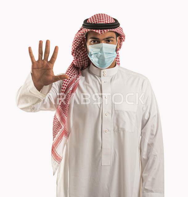الحفاظ على المسافات وعدم الملامسه السطحيه، إتباع الاحترازات الوقائيه ضد كوفيد 19 ، بورتريه لرجل عربي خليجي سعودي يرتدي الثوب والزي التقليدي يضع القناع الطبي الوقائي للوقايه من فايروس كورونا يرفع يده لتجنب المصافحة واقتراب الاخرين منه، خلفيه بيضاء