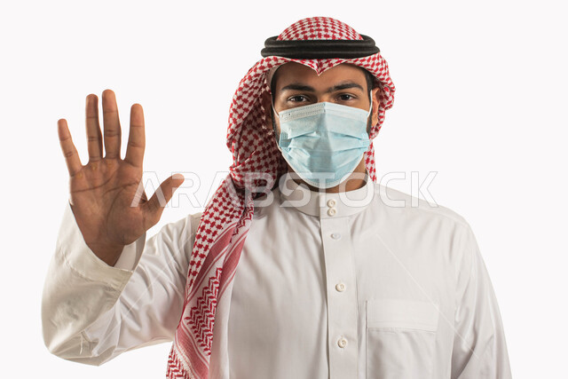 إتباع  الاحترازات الوقائيه ضد كوفيد 19 ، بورتريه مقرب لرجل عربي خليجي سعودي يرتدي الثوب والزي التقليدي يضع القناع الطبي الوقائي للوقايه من فايروس كورونا يرفع يده لتجنب المصافحة واقتراب الاخرين منه ،الحفاظ على المسافات وعدم الملامسه السطحيه، خلفيه بيضاء