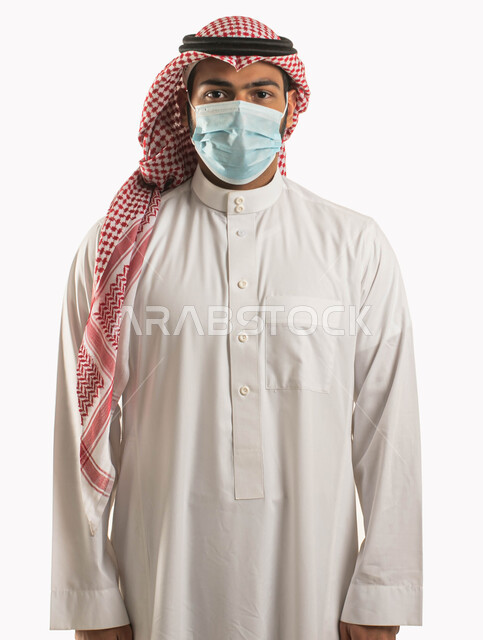 إتباع الإجراءات و الاحترازات الوقائيه ضد كوفيد 19 ، بورتريه مقرب لرجل عربي خليجي سعودي يرتدي الثوب والزي التقليدي يضع القناع الطبي الوقائي للوقايه من فايروس كورونا وينظر إلى الكاميرا ، الحفاظ على المسافات وتجنب الملامسه السطحيه، خلفيه بيضاء