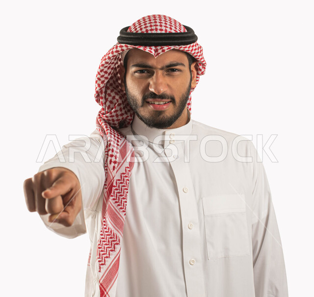 إيماءات وجه ويدين تدل على الإتهام وإلقاء اللوم، بورتريه لمواطن عربي خليجي سعودي يرتدي الشماغ والثوب التقليدي ويشير بأصبع السبابة بغضب وعبوس، الشعور بالإنزعاج والرغبة بالإنتقام، خلفية بيضاء