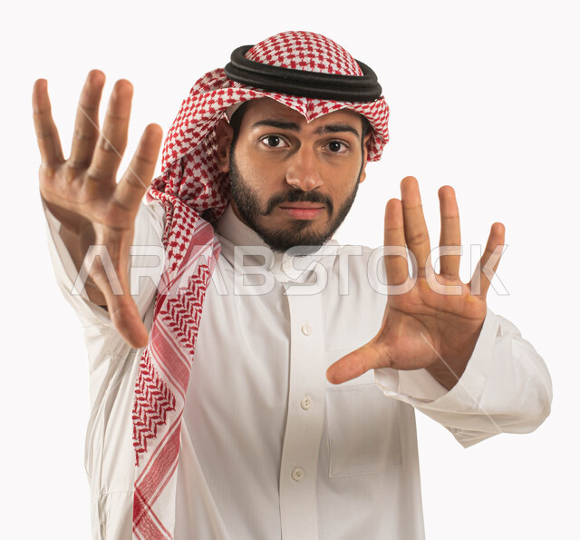 مفهوم الرعب والذعر والرهبة، بورتريه لشاب عربي خليجي سعودي يرتدي الشماغ والثوب التقليدي ويرفع يده لأعلى بإيماءات الخوف والقلق، تعابير واشارات لطلب التوقف والإمتناع عن فعل شيء ما، خلفية بيضاء