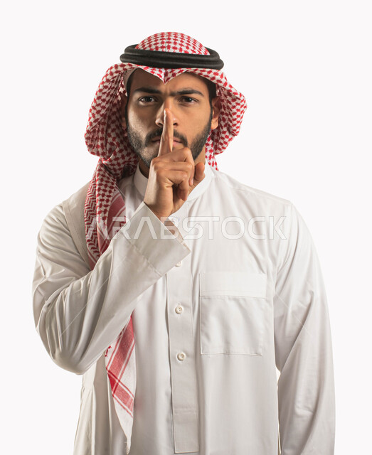 Stop talking and lower the volume, a portrait of a Saudi Gulf Arab man wearing a shemagh and a traditional dress and putting his index finger on his mouth with angry facial gestures, calm and stillness and do not disturb, signals requesting silence and silence, white background
