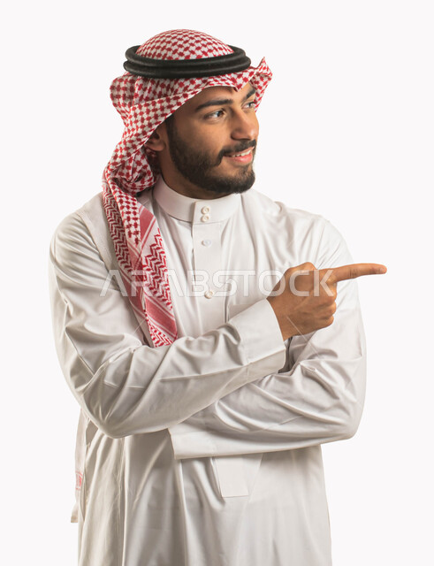 ايماءات السرور والسعادة، بورتريه لرجل عربي خليجي سعودي مبتسم يرتدي الشماغ والثوب التقليدي ويقف بيد مكتوفة وينظر الى مكان ما، الاشارة بأصبع السبابة نحو شيء وتحديد هدف ما، خلفية بيضاء
