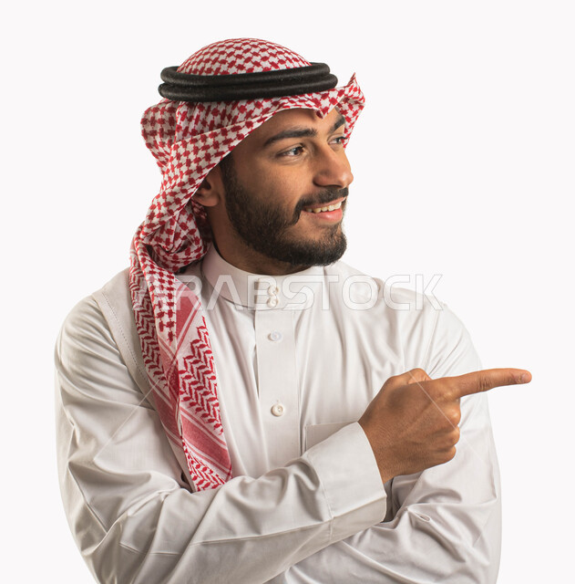 الاشارة بأصبع السبابة نحو شيء وتحديد هدف ما، بورتريه لرجل عربي خليجي سعودي مبتسم يرتدي الشماغ والثوب التقليدي ويقف بيد مكتوفة وينظر الى مكان ما، ايماءات السرور والسعادة، خلفية بيضاء