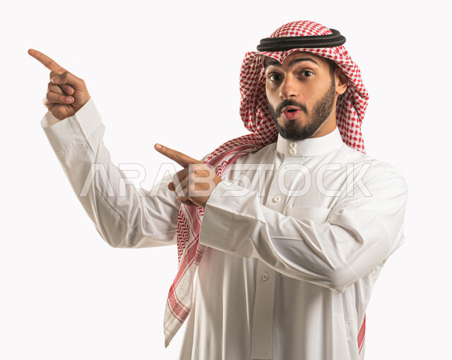 الصدمة والتعجب من شيء ما، بورتريه لرجل عربي خليجي سعودي يرتدي الشماغ والثوب التقليدي ويشير بأصبع السبابة نحو مكان لتحديد هدف ما، النظر الى الكاميرا بايماءات وجه تدل على الذهول والدهشة، خلفية بيضاء