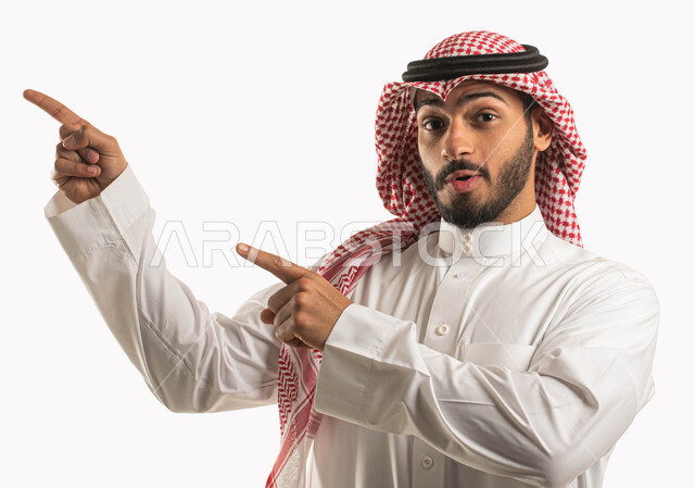 النظر الى الكاميرا بايماءات وجه تدل على الذهول والدهشة، الصدمة والتعجب من شيء ما، بورتريه لرجل عربي خليجي سعودي يرتدي الشماغ والثوب التقليدي ويشير بأصبع السبابة نحو مكان لتحديد هدف ما، خلفية بيضاء