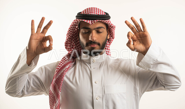 ممارسة رياضة اليوغا التأملية، بورتريه لرجل عربي خليجي سعوي يرتدي الثوب التقليدي والشماغ ويغمض عينيه ويرفع يديه إلى أعلى بإيماءات وتعابير التأمل والاسترخاء، أنشطة وتمارين رياضية، التنفس العميق والتركيز، خلفية بيضاء