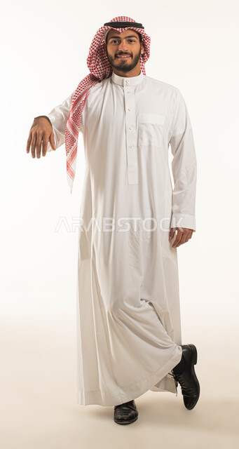 حركة يد واشارات لطلب القبلة لتنفيذ امر ما، بورتريه لشاب وسيم عربي خليجي سعوي يرتدي الشماغ والثوب التقليدي ويقف بإستقامة وينظر الى الكاميرا بسرور وسعادة، ايماءات الاستناد على رجل واحدة، الارتكاز على شيء ما، صورة الجسم بالطول الكامل، خلفية بيضاء
