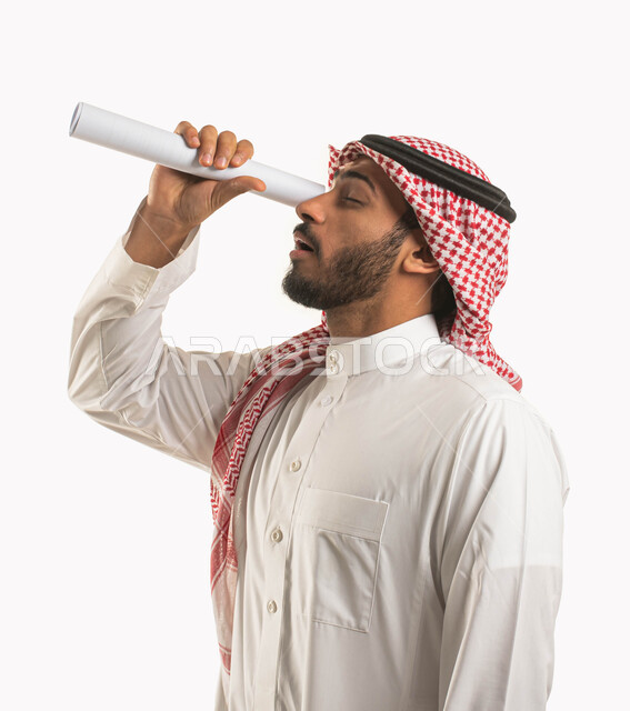 النظر إلى شيء ما, بورتريه من الجانب لشاب عربي سعودي خليجي يرتدي الثوب و الشماغ التقليدي ينظر من خلال ورقة بيضاء ملفوفة, التظاهر باستخدام المايكروسكوب, التخطيط للمستقبل البعيد, تقريب الأشياء البعيدة, إيماءات التركيز و التدقيق لرؤية شيء ما, خلفية بيضاء .