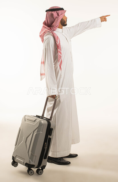رحلة عمل في الخارج, الاستعداد للسفر في جولة سياحية ممتعة, بورتريه من الجانب لشاب عربي سعودي خليجي يرتدي الثوب و الشماغ التقليدي يمسك حقيبة سفر و يشير إلى مكان ما, التسلية و الترفية في العطلة الصيفية, صورة الجسم بالطول الكامل, خلفية بيضاء .