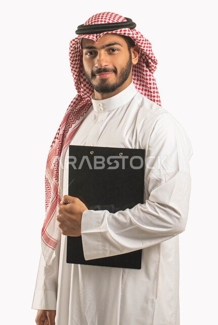 كتابة المهام و الأهداف اليومية, بورتريه لرجل أعمال عربي سعودي خليجي مبتسم يرتدي الثوب و الشماغ التقليدي يمسك بيده سنادة ورق سوداء, قرطاسية و أغراض مكتبية, إيماءات الثقة و النظر إلى الكاميرا, خلفية بيضاء .
