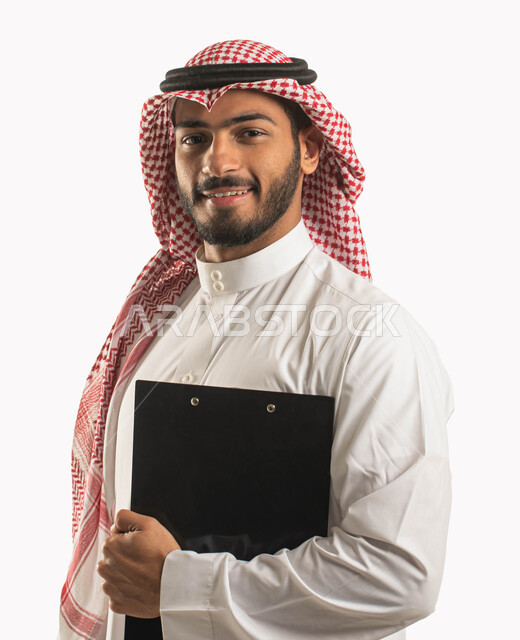أدوات تدوين المهام و الملاحظات, بورتريه مقرب لرجل أعمال عربي سعودي خليجي مبتسم يرتدي الثوب و الشماغ التقليدي يمسك بيده سنادة ورق سوداء, قرطاسية و أغراض مكتبية, إيماءات الثقة و النظر إلى الكاميرا, خلفية بيضاء .