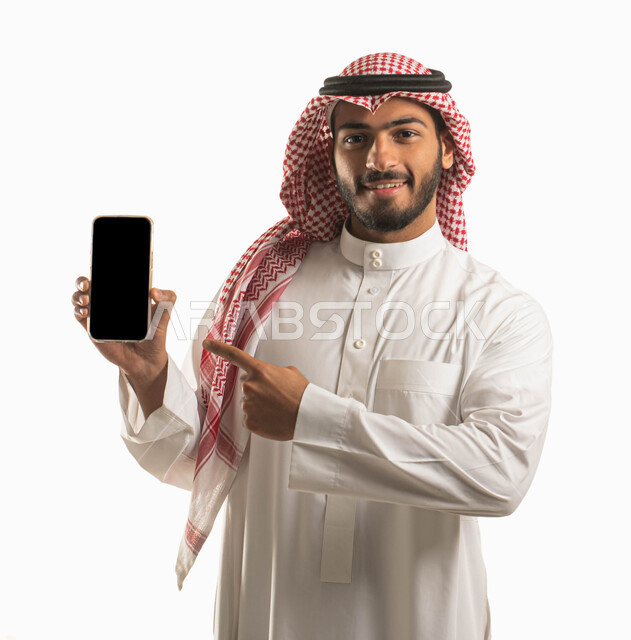 استخدام التكنولوجيا المتطورة للتسويق الرقمي الإلكتروني، عرض تطبيقات و خدمات الهاتف المحمول، بورتريه لشاب عربي سعودي  خليجي مبتسم يرتدي الشماغ و الثوب التقليدي يشير باصبعه إلى جهاز الجوال بشاشة سوداء فارغة، برامج التواصل الاجتماعي عبر الهاتف, خلفية بيضاء .