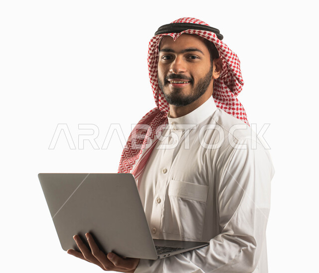 تصفح الإنترنت و إنجاز المهام عبر اللابتوب, بورتريه مقرب لرجل مبتسم عربي سعودي خليجي يرتدي الثوب والشماغ التقليدي يحمل بيده جهاز الحاسب المحمول, إستخدام أجهزة تقنية حديثة, احتراف وظائف الحاسوب و تكنولوجيا المعلومات, خلفية بيضاء .