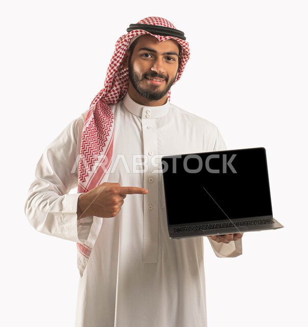 احتراف وظائف الحاسوب و تكنولوجيا المعلومات, بورتريه لرجل مبتسم عربي سعودي خليجي يرتدي الثوب والشماغ التقليدي يحمل بيده جهاز الحاسوب المحمول ويشير إليه, تصفح الإنترنت و إنجاز المهام عبر جهاز اللابتوب, عرض شاشة سوداء فارغة, مفهوم التسويق الألكتروني, خلفية بيضاء .