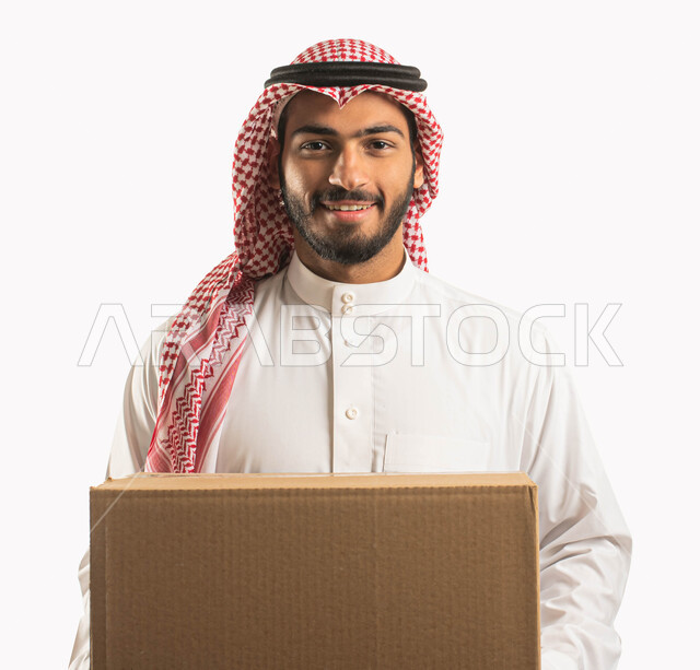 خدمات الشحن و التوصيل ذات الجودة العالية، التسوق وطلب المنتجات عبر الإنترنت، عملية توصيل الطلبات للزبائن، بورتريه مقرب لمندوب عربي سعودي خليجي يرتدي الشماغ والثوب التقليدي ويحمل بيده صندوق كرتوني، خلفية بيضاء
