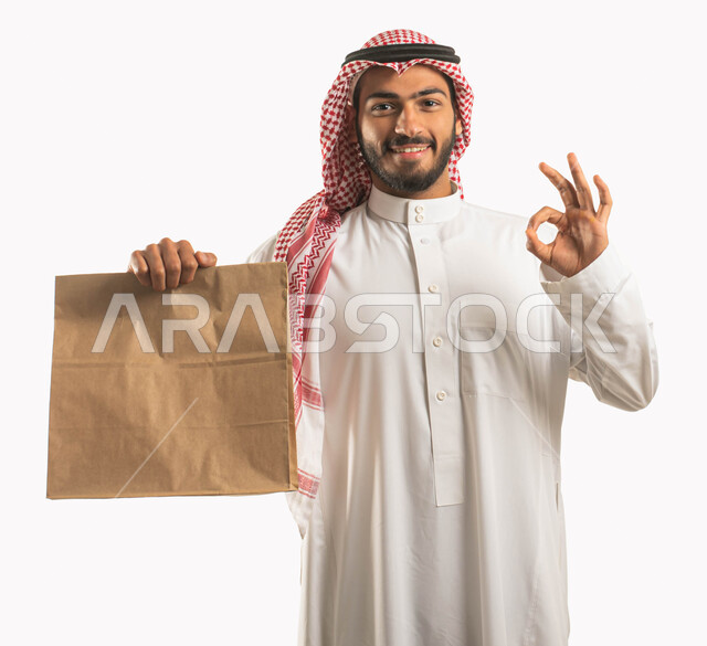 Gestures of happiness and pleasure with the arrival of purchases, high-quality shipping and delivery services, a portrait of a Saudi Gulf Arab man wearing a shemagh and traditional thobe holding a delivery package in his hand, shopping and ordering products online and delivering them to customers, a sign of quality, acceptance and approval, white background.