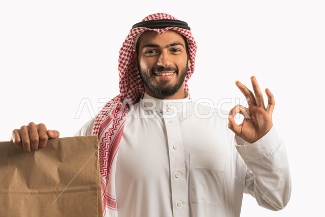 خدمات الشحن والتوصيل ذات الجودة العالية، بورتريه مقرب لرجل عربي سعودي خليجي يرتدي الشماغ والثوب التقليدي  يحمل بيده طرد التسليم، التسوق وطلب المنتجات عبر الإنترنت وتوصيلها للزبائن، ايماءات السعادة والسرور بوصول المشتريات، علامة الجودة والقبول والموافقة، خلفية بيضاء