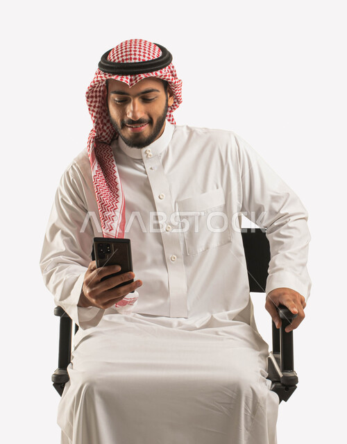 تصفح وسائل التواصل الاجتماعي، ايماءات الاندماج والتركيز، بورتريه مقرب لرجل عربي سعودي خليجي مبتسم يرتدي الشماغ و الثوب التقليدي يجلس على كرسي أسود مريح ويستخدم الهاتف المحمول,، التحدث والدردشة مع الأصدقاء عبر الجوال، مفهوم الراحه والجلوس ,  خلفية بيضاء
