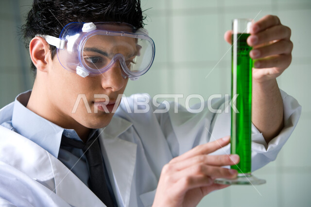 القيام بالأنشطة التعليمية والتجارب في بيئة عملية متطورة, البحث والتطوير وإجراء التفاعلات الكيميائية, طالب جامعي عربي خليجي إماراتي يقوم بتجربة علمية في المختبر, ارتداء نظارة الوقاية والمعطف الأبيض, اكتساب المعرفة والخبرة بإيماءات الدقة والتركيز في التجربة